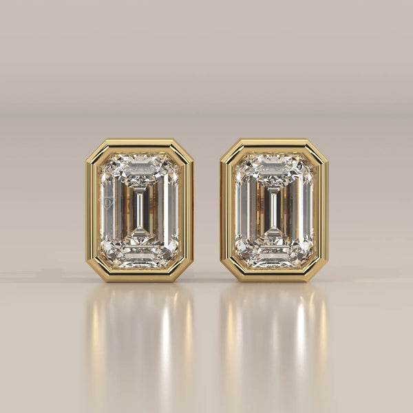 A pair of emerald-cut diamond stud earrings in a yellow gold bezel setting.