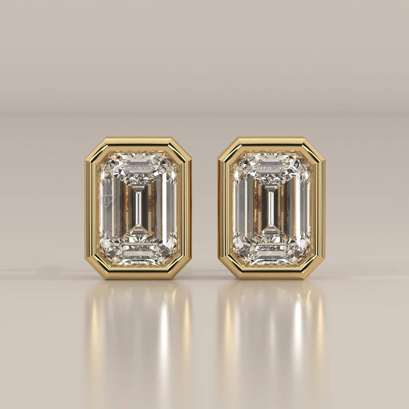 A pair of emerald-cut diamond stud earrings in a yellow gold bezel setting.