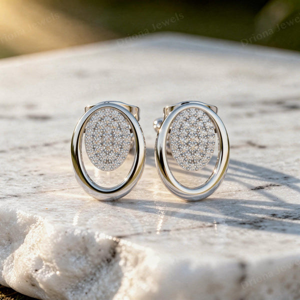 Oval pave diamond stud earrings in a solid gold open-frame design on a white stone surface.